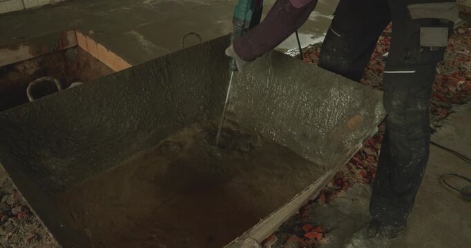 Professional builder using an electric mixer to prepare a fresh cement solution in a large container at a construction site, getting ready for floor screeding and house renovation