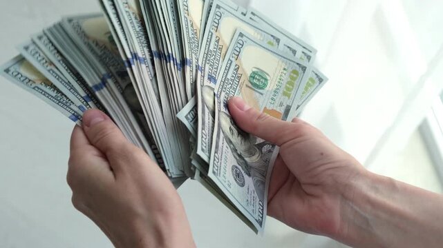 Hands hold stacks of U.S. Currency. Fingers gently fan out bills for counting. Bright background highlights the money's crisp edges.