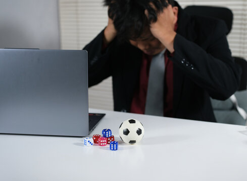 A stressed man loses in online soccer gambling with dice, a soccer ball, and a laptop on the table. A gambler loses a lot of money playing soccer, a gambling addiction. Loss, ruin, debt, the concept o