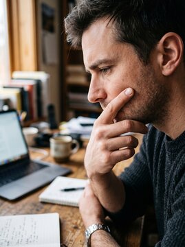 A contemplative young man sitting at a rustic wooden desk with a laptop, open notebook, and coffee mug, reflecting deeply while working in a cozy, well-lit home office environment.