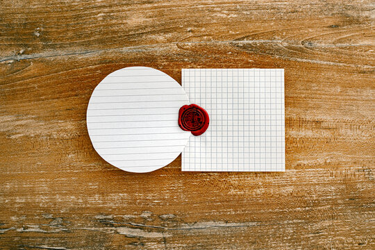 Red wax seal stamping together a round lined paper and a square graph paper on a rustic wooden table. Conceptual metaphor for official agreement, contract validation, or business alliance.
