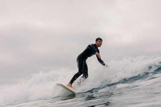 A man in a black wetsuit rides a wave under an overcast sky.