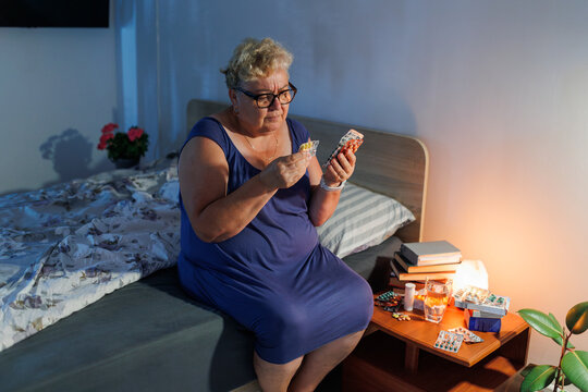 Overweight mature woman with glasses reading medical pills in bedroom.