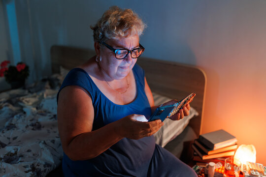 Mature female holding medical pills and smartphone in dark bedroom during night.
