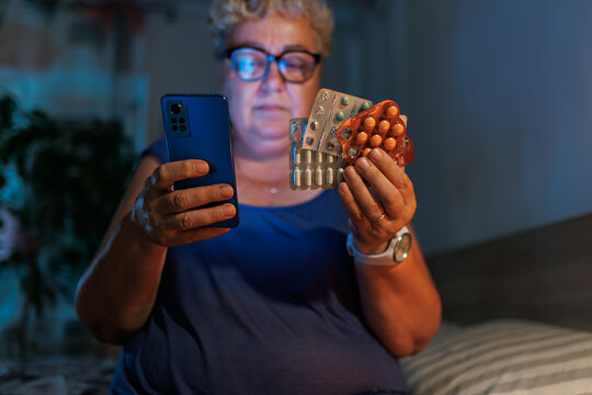 Female reading smartphone holding medical pills in dark bedroom late at night.