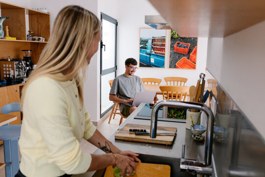 A woman in a yellow cardigan cooks while a man sits at a laptop.