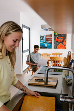 A woman works at the sink while a man uses a laptop nearby.