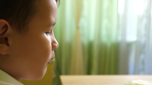 Boy eats corn puffs while sitting at the table, close side profile in a cozy home setting with natural light.