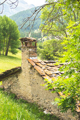 An old, rustic stone cottage with a textured slate-tile roof and a prominent stone chimney sits nestled on a green hillside framed by tree branches. Hameau de Maljasset, Saint Paul sur Ubaye, Haute, A © pauline.mongarny