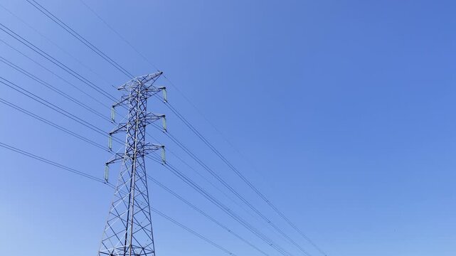 High-voltage tower stands tall against vivid blue sky. Thin lines stretch across the frame, guiding power through the air. One small cloud drifts gently, adding calm to the scene.
