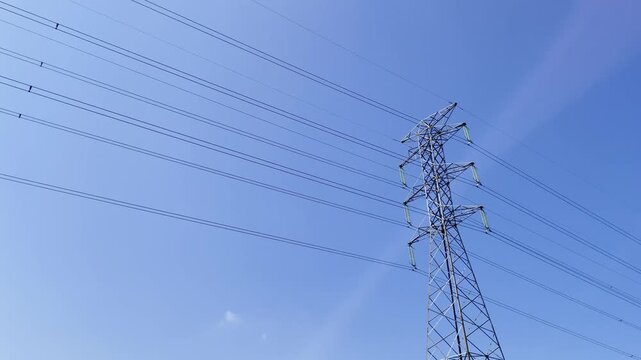 High-voltage tower stands tall against vivid blue sky. Thin lines stretch across the frame, guiding power through the air. One small cloud drifts gently, adding calm to the scene.