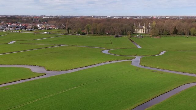 Aerial medium shot of a scenic Dutch landscape with winding drainage canals flowing through lush green polder fields toward a historic estate and castle nestled in a dense forest
