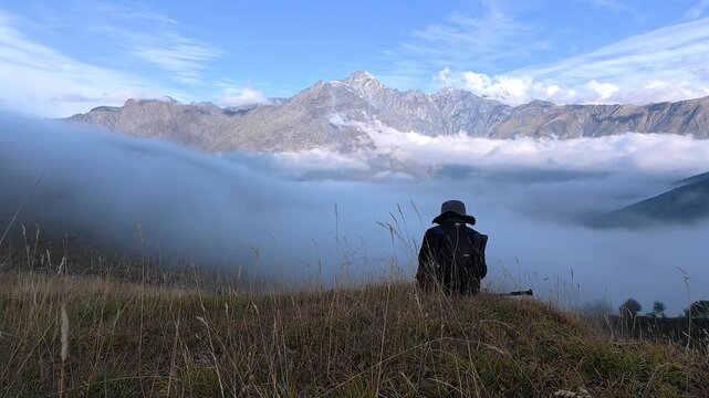 4K real time video of a solo hiker sitting on a grassy hill looking at thick white fog over mountains.
