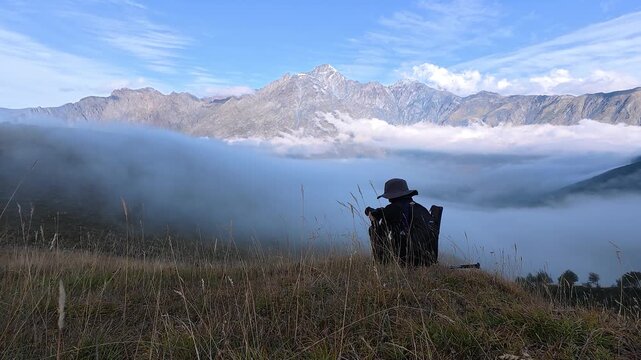 4K timelapse video of a solo traveler sitting on a grassy hill looking down at thick low clouds moving through the mountain valley.