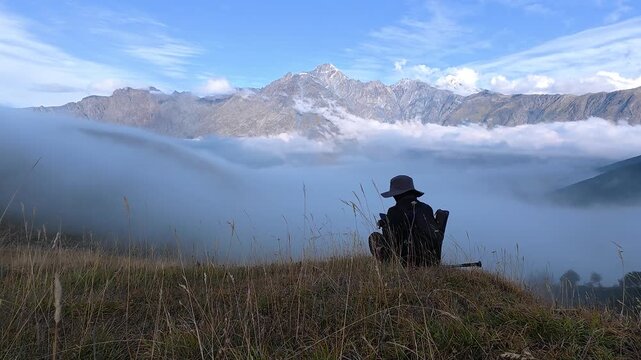 4K timelapse video of a solo hiker sitting on a grassy mountain hill watching thick white fog moving over the peak.