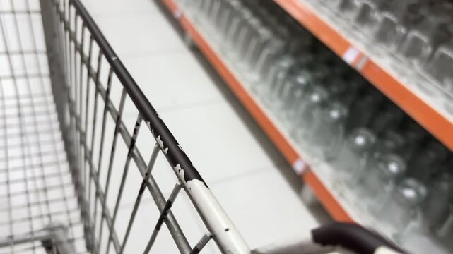 POV shot of empty shopping trolley moving past glass jars and cups on display shelves in grocery store.