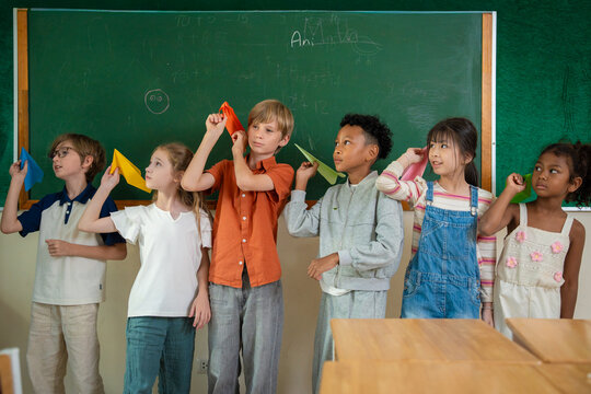 Diverse elementary school children standing in front of chalkboard holding colorful paper airplanes. Multiethnic primary students presenting their origami paper planes during creativity in classroom.