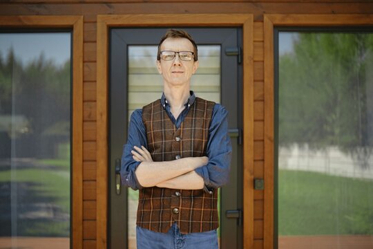 Middle-aged man in blue shirt and brown vest stands confidently in front of modern wooden house doors