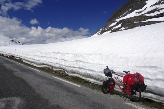 A bicycle with red panniers stands next to a snow-covered mountain road under a clear sky, Fjell in spring, Geiranger, Langevatn, Norway