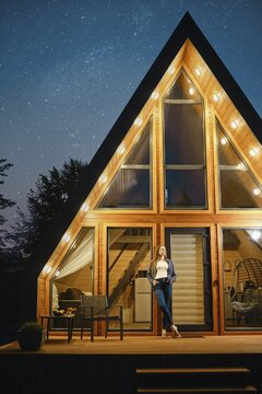 Dreamy woman stands on the doorway of a large wooden cabin late at night