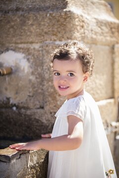 Little girl in white dress stands by a fountain, La Lastra del Cano, Spain