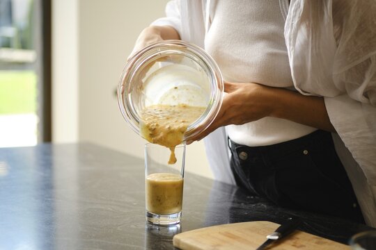 Pouring a creamy banana and sea buckthorn smoothie from a glass bowl into a tall glass. The setting is a bright kitchen with a sleek countertop and a cutting board nearby