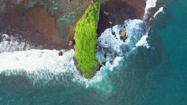 Aerial drone footage on top of Watulawang island in Ngandong beach, Gunungkidul regency, Wonosari, Java island, Indonesia, with green grasses, blue sea water, and waves crashing reef