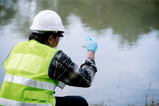 Ecologist examining water sample using a flask while standing by the riverbank wearing protective gear and a safety vest