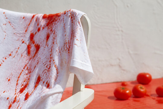 A white t-shirt stained with red liquid is draped over a chair against a white textured wall with some red objects on the floor.