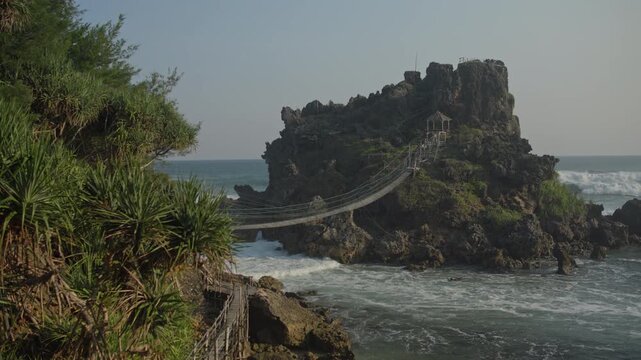 A suspension rope bridge crosses turbulent surf to a rocky island shrine perched on dramatic cliffs, framed by lush tropical vegetation and rugged shoreline under soft, natural daylight.