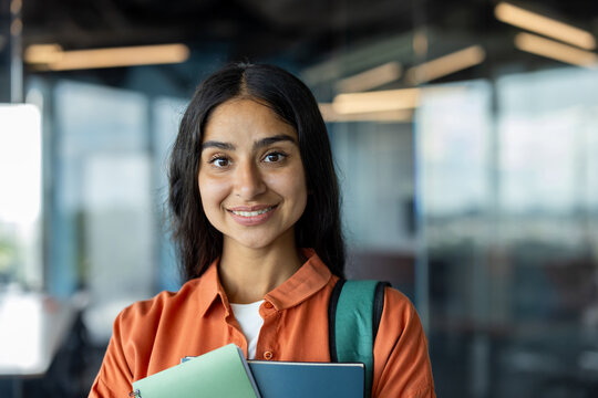 Close-up portrait of a young Indian female student standing in a university building, holding a backpack and books, smiling at the camera