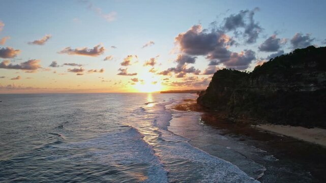 Aerial drone footage of Watunene beach edge in Gunungkidul regency, Yogyakarta area, Java island, Indonesia, with high cliff silhouette,  turquoise water, waves , at sunset, facing the sun with clouds