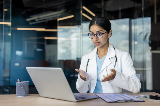 Indian young female doctor sitting at office desk in hospital and talking on video call on laptop camera