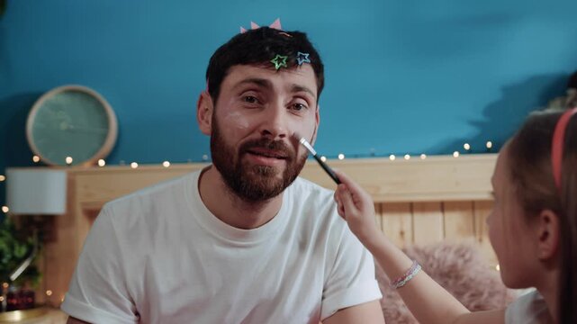 Portrait of poor tired father playing with his daughter at home. Side view little daughter applying cosmetics on her father's face having fun in bedroom.