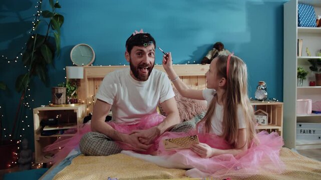 Fun family scene at party. Portrait of handsome man playing the ape crying for help and laughing. His little sweet daughter applying cosmetics on face making him a pretty princess.