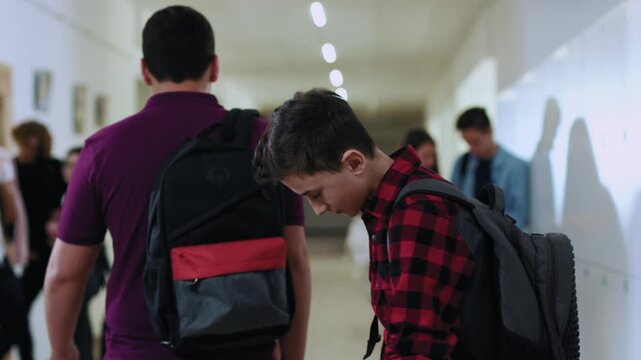 Cute schoolboy with backpack and books standing in school corridor. His rude classmate pushes him to the side. Embarassed young boy upset with the situation. College and people concept.