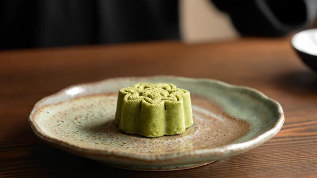 Hands placing green matcha mooncake with intricate floral pattern on rustic plate