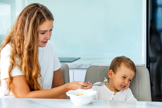 Mother unsuccessfully trying to feed child in the kitchen, little son refusing food that mom gives. Child nutrition, conflicts, parenting, family, health, childcare products, pediatric healthcare.