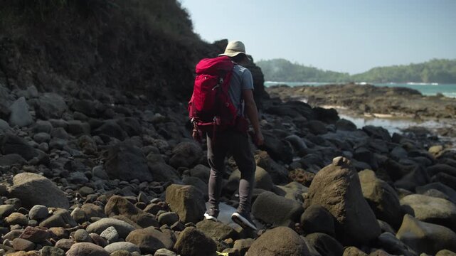 Solo backpacker with a red backpack walking across rocky beach cliffs on a bright morning, exploring a tropical coastal landscape during an outdoor travel adventure