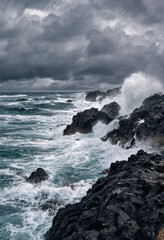 Fototapeta premium Powerful crashing ocean waves hitting dark volcanic rocks under a dramatic stormy dark sky