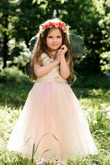 Fototapeta premium Lovely little girl wearing a flower wreath and a pink tulle dress posing with hands folded near her face in a sunny summer forest.