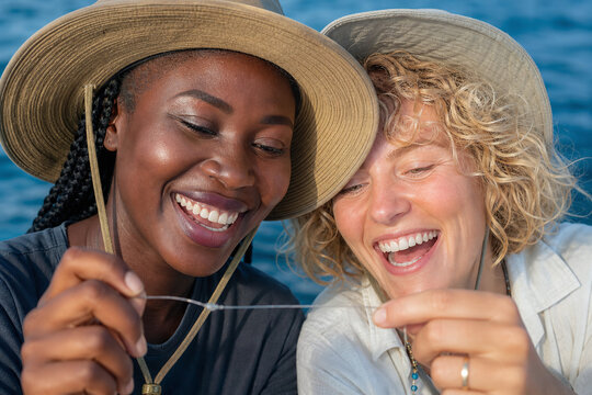 Jolly Malian and English blonde women friends checking a knot on a fishing boat in glowing sunlight &ndash; teamwork, bonding, maritime activity, friendship, multicultural, boat life, commercial stock