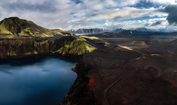 Aerial view of Blahylur crater lake surrounded by vibrant green mossy slopes and dark volcanic terrain with a winding dirt road, Rangarthing ytra, Iceland.