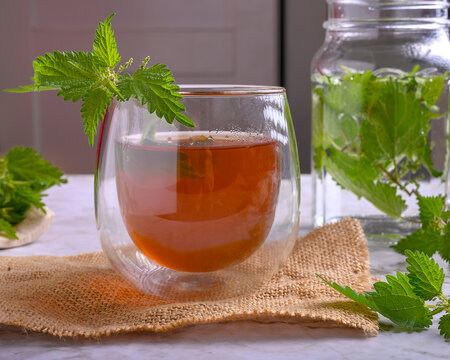 Nettle herbal tea in a double wall glass on burlap and marble surface, fresh stinging nettle leaves around a jar with water, concept of healthy detox beverage and natural medicine