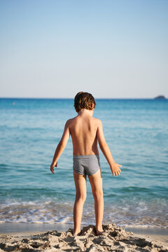 A child wearing striped swim trunks stands on the sand, gazing out at the beautiful turquoise water during a summer vacation.