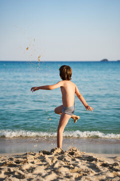 A carefree child in swim trunks kicks and tosses sand into the air near the ocean shoreline, enjoying outdoor recreation and freedom on a coastal holiday.
