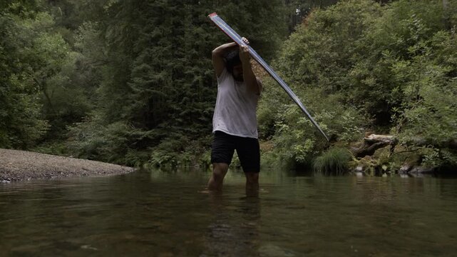 Man Swinging Board Across Shallow Stream Surface and Making Splash In Slow Motion - Low, Wide Shot