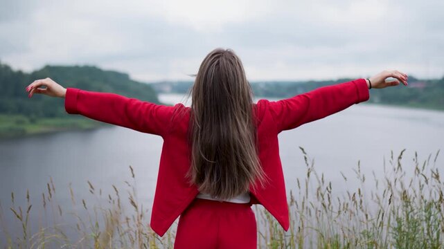 Young woman in a red outfit stands by a calm lake with arms wide open, feeling freedom, strength, lightness and connection with nature, like a flying bird