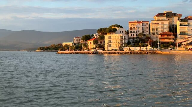 Scenic coastal view of Gulluk, Turkey, featuring white resort hotels and residential buildings along the waterfront, illuminated by the warm glow of late afternoon sun.