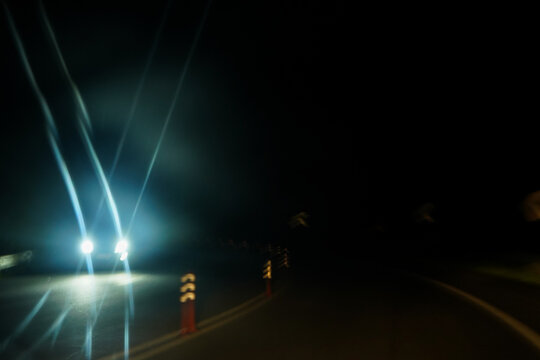 Blinding car headlights on a dark night road bend with reflective traffic markers. Abstract long exposure motion blur of vehicle high beams on a sharp highway curve.